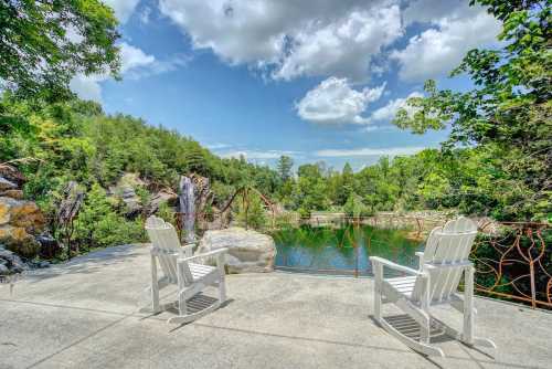 Two white Adirondack chairs overlook a serene pond surrounded by lush greenery and a bright blue sky with clouds.