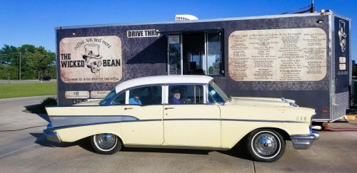 A vintage yellow car parked next to a food truck named "The Wicked Bean," featuring a skull logo and menu.