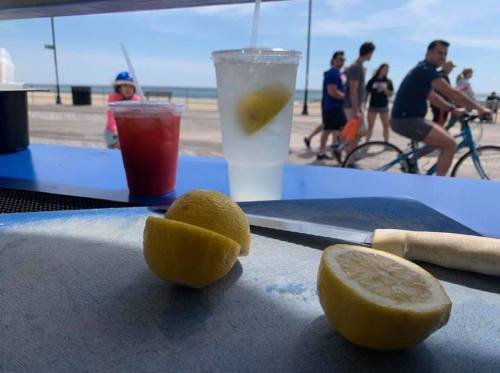 Lemons on a cutting board with drinks in the background, beachgoers and cyclists enjoying a sunny day by the shore.