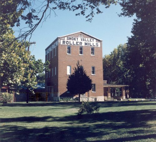 Historic Smoky Valley Roller Mills building surrounded by trees and a grassy area under a clear blue sky.