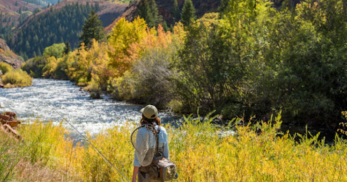 A person stands by a river, surrounded by colorful autumn foliage and mountains in the background.