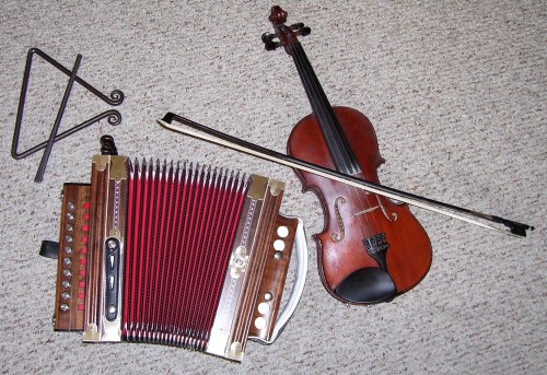 A violin, an accordion with red and wooden details, and a triangle resting on a textured surface.