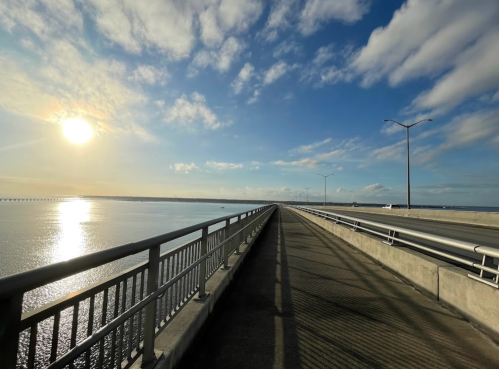 A wide bridge stretches over calm water, with a bright sun and blue sky in the background.