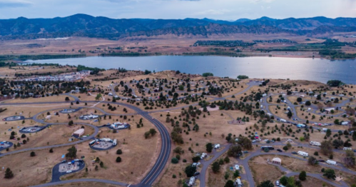 Aerial view of a lakeside area with winding roads, campsites, and mountains in the background under a cloudy sky.