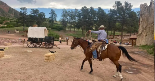 A cowboy on horseback holds a lasso, with a wagon and people in a rustic outdoor setting surrounded by trees.