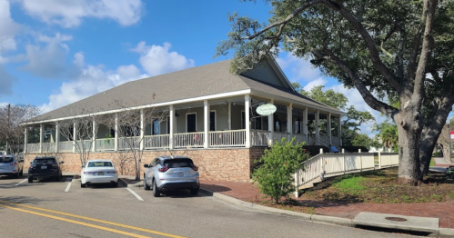 A large, light-colored building with a porch, surrounded by trees and parked cars on a sunny day.