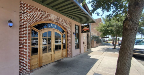 Exterior view of a brick building with wooden double doors, street trees, and a sunny sidewalk. Address: 1308.