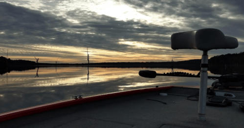 A serene lake at sunset, with clouds reflecting on the water and a boat's seat in the foreground.
