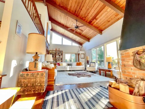 Bright, spacious living room with wooden beams, large windows, cozy furniture, and a fireplace.