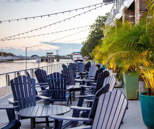 Outdoor seating area with dark wooden chairs and tables, string lights overhead, by a waterfront at sunset.
