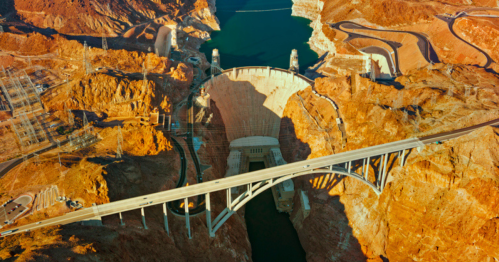 Aerial view of a large dam surrounded by rocky terrain and a river, with a bridge crossing above.