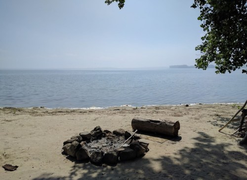 A serene beach scene with a calm water view, a fire pit made of stones, and a log on the sandy shore.