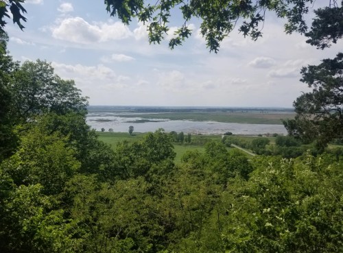 A scenic view of a lush green landscape with a river and open fields under a partly cloudy sky.