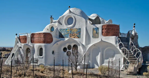 Whimsical white house with curved shapes, colorful murals, and unique architectural features against a clear blue sky.
