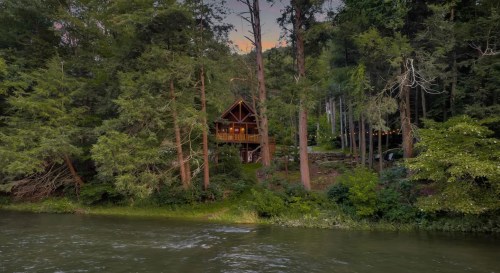 A cozy cabin nestled among tall trees by a serene river at dusk, with warm lights glowing from the windows.