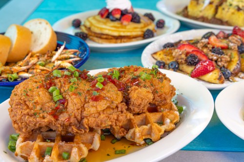A plate of fried chicken and waffles drizzled with syrup, surrounded by various desserts and sides on a colorful table.