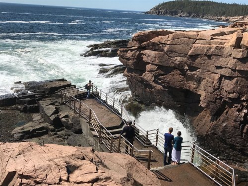 Two people stand on a viewing platform by the ocean, with waves crashing against the rocks in a scenic coastal landscape.