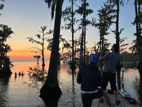 Two people on a boat photographing a sunset over a serene lake surrounded by cypress trees.