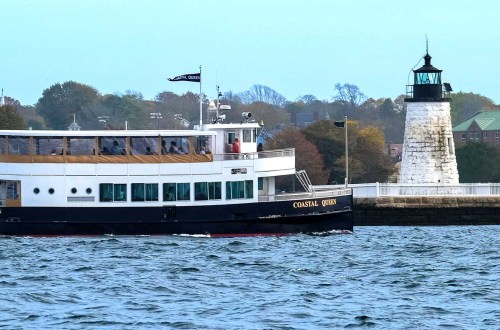 A boat named "Coastal Queen" sails past a lighthouse on a calm waterway, with trees in the background.