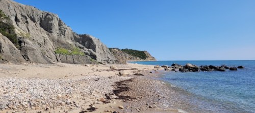 A serene beach scene with rocky cliffs, clear blue water, and a sandy shore under a bright blue sky.