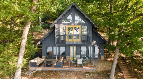 A modern A-frame cabin surrounded by trees, featuring large windows and a deck with outdoor seating.