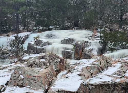 A frozen waterfall cascades over rocky terrain, surrounded by snow and trees in a winter landscape.