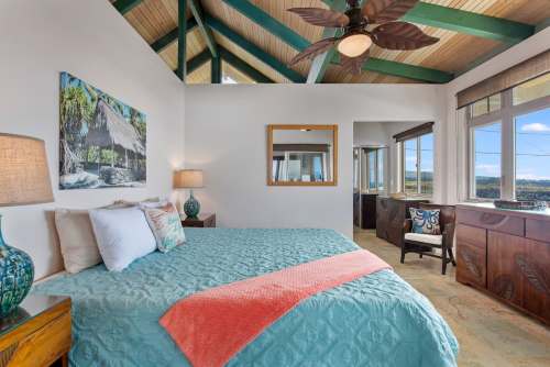 Bright bedroom with a high ceiling, featuring a turquoise bedspread, tropical decor, and large windows with scenic views.