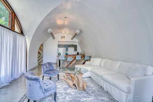 Modern living room with a curved ceiling, white sofa, two blue chairs, and a wooden coffee table. Bright and airy atmosphere.