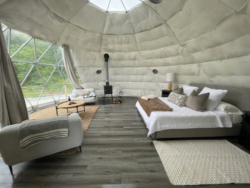 Cozy, modern geodesic dome interior with a bed, seating area, and large windows overlooking nature.