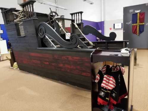 A wooden pirate ship play structure with a steering wheel and pirate costumes hanging nearby.