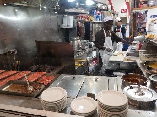 A busy kitchen with a chef preparing food, hot dogs on the grill, and plates stacked on the counter.
