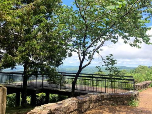 A scenic overlook with a wooden deck surrounded by trees, offering a view of rolling hills and a blue sky.