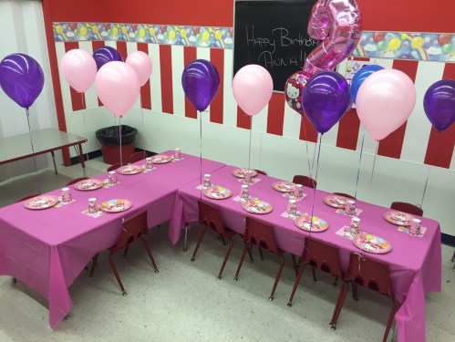 A festive birthday setup with pink and purple balloons, decorated tables, and plates for a celebration.