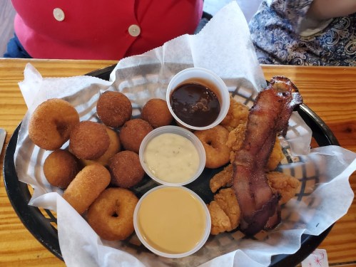 A basket of fried food featuring small doughnuts, chicken strips, bacon, and three dipping sauces.