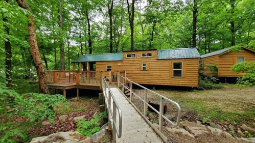 A wooden cabin surrounded by trees, featuring a ramp and a deck, nestled in a lush green forest.