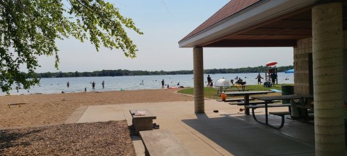 A beach scene with people swimming and relaxing by the water, viewed from a shaded pavilion.