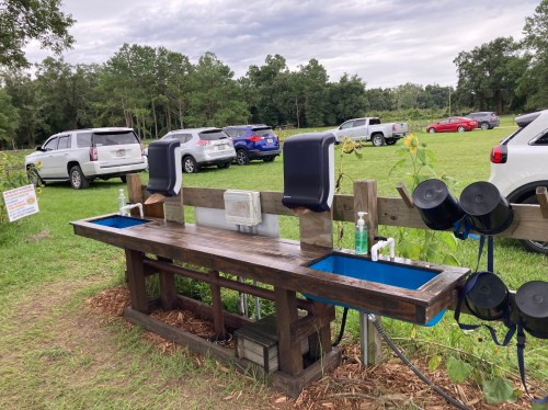 Outdoor handwashing station with two sinks, soap dispensers, and a row of parked cars in the background.