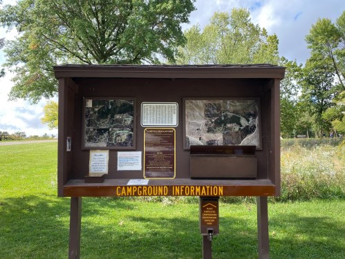 A wooden information kiosk displaying maps and details about a campground, set in a grassy area with trees.