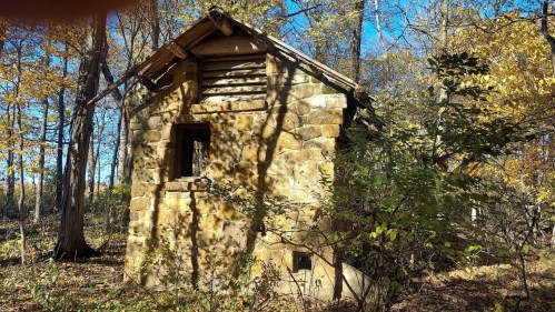 A small, weathered stone building surrounded by trees with autumn leaves, under a clear blue sky.