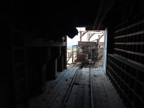 Interior of an abandoned building with wooden floors, tracks, and a view of a structure outside.