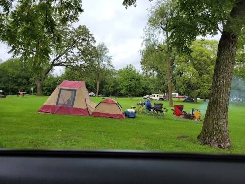 A campsite with a tent, chairs, and a cooler on green grass, surrounded by trees and a cloudy sky.