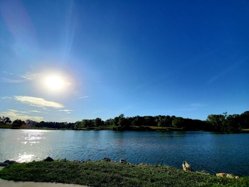 A serene lake under a clear blue sky, with the sun shining brightly and lush greenery in the background.