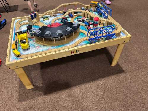 A wooden train table with tracks, vehicles, and playsets, set on a carpeted floor.