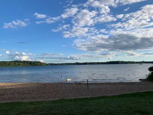 A serene lake view with sandy shore, gentle waves, and a blue sky dotted with clouds.