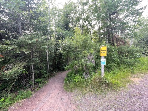 A narrow dirt path leads into a lush green forest, marked by a yellow sign for a state park.