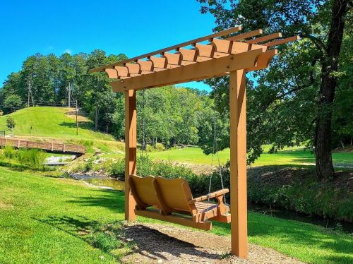 A wooden swing under a pergola in a green park, surrounded by trees and a small creek. Clear blue sky above.