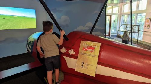 A child climbs on a red airplane exhibit in a museum, with a screen showing a landscape in the background.