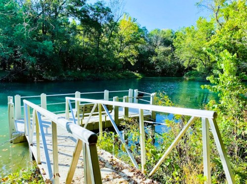 A serene dock extends over a clear blue river, surrounded by lush green trees and a bright blue sky.