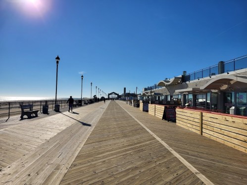 A sunny boardwalk with wooden planks, lined by benches and shops, leading to the beach under a clear blue sky.