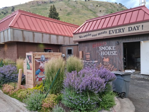 Exterior of Bubba's Smoke House with a red roof, surrounded by greenery and lavender, featuring a sign that says "We smoke our meats EVERY DAY!"